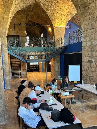 A group of people sit at tables in a vaulted stone hall with a flip chart, a projector and a spiral staircase in the background.