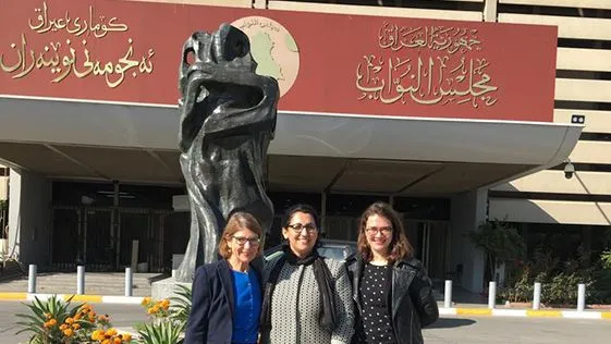 Three women standing in front of a building entrance with a statue and a red sign bearing Arabic and Kurdish script.