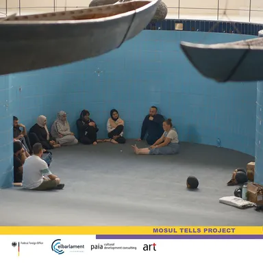 A group of people sitting in a circle on the floor of a round blue-tiled room with sculptural mask objects hanging overhead, above a "MOSUL TELLS PROJECT" banner.