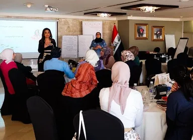 A seated audience of mostly women wearing headscarves in a conference room, with two women presenting at the front near an Iraqi flag.