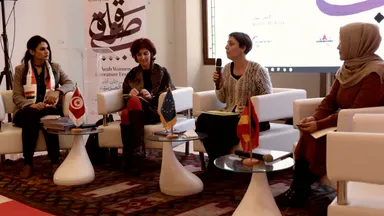 Four women seated on a panel, one speaking into a microphone, in front of a banner reading "Arab Women Literature Festival" with small national flags on tables beside them.