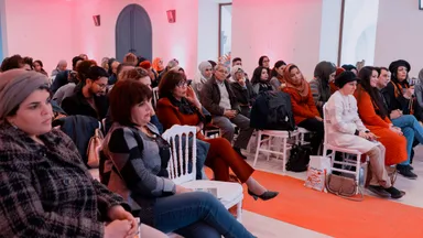 An audience seated in rows in a room lit pink, most attendees wearing headscarves, listening attentively.