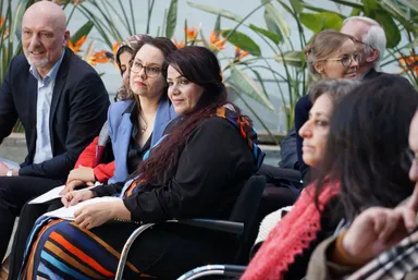 A group of people sit outdoors in front of tropical plants and water, one of them in a wheelchair, watching something off-camera.