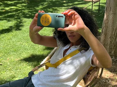 A woman sits in a wicker chair outdoors on grass, holding up a smartphone with a yellow sticker on its back as if taking a photo.