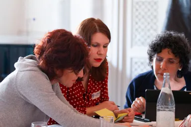 Three women sit close together at a table with a laptop and a glass bottle, looking down at papers.