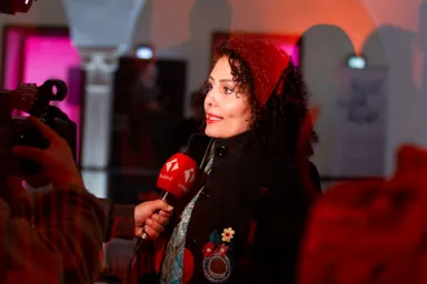 A woman with curly hair and a red headscarf is interviewed by an unseen person holding a red microphone with a logo and Arabic text, in a venue with pink lighting.
