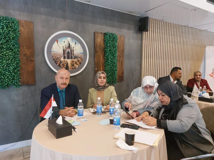 A standing woman wearing a headscarf gestures while speaking, in front of four seated women listening.