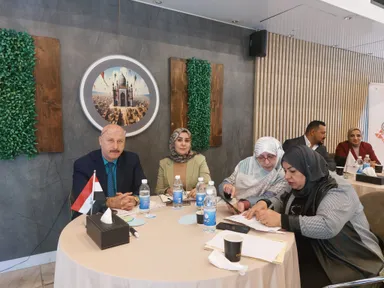 A standing woman wearing a headscarf gestures while speaking, in front of four seated women listening.