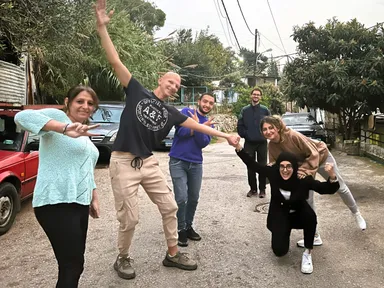 A group of people posing playfully with raised arms and peace signs on a street lined with parked cars and trees.