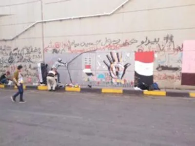 A person walks along a street past a wall covered in graffiti and a mural of a raised hand and the Iraqi flag, with yellow curb markings and people nearby.