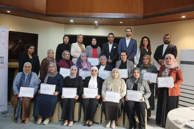 A group of people, mostly women in headscarves, pose indoors holding certificates in front of a partially visible banner.