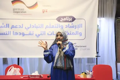 A woman in a blue robe speaks into a microphone in front of a banner with Arabic text and the logos "german cooperation", "giz" and "elbarlament".