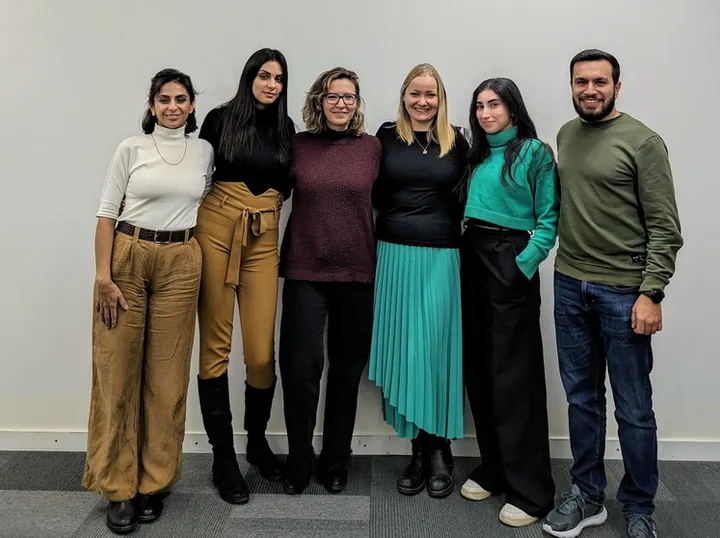 A group of six people stand arm in arm against a plain wall, smiling for a group photo.