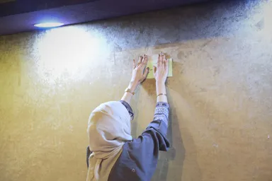A person presses a sticky note onto a wall, seen from behind with hands raised and wearing bracelets.